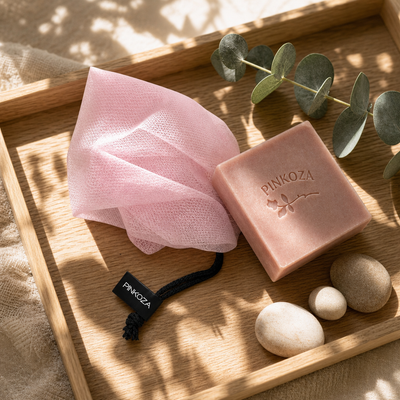 PINKOZA pink mesh soap bag with a black logo tag resting on a wooden tray next to a pink goat milk soap bar, eucalyptus leaves, and smooth stones.